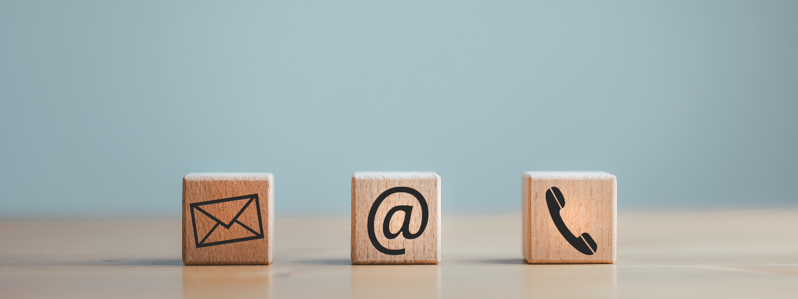 Contact us icons in wood. Mail on the left, email in the center, and phone on the right on a table in a blue room.