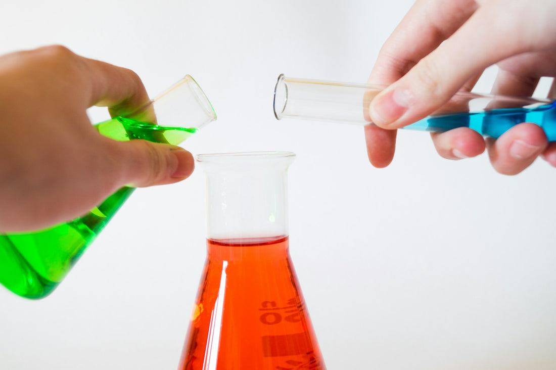 Three chemicals, green, blue, and red. Blue and green are being poured into the red beaker.