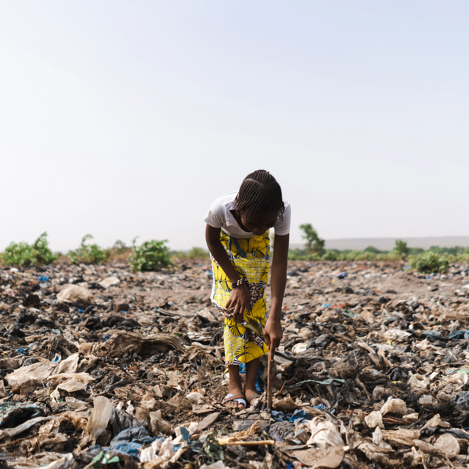 A young child standing in the center of plastic pollution in her environment. She pokes the towering pile of waste with a stick.