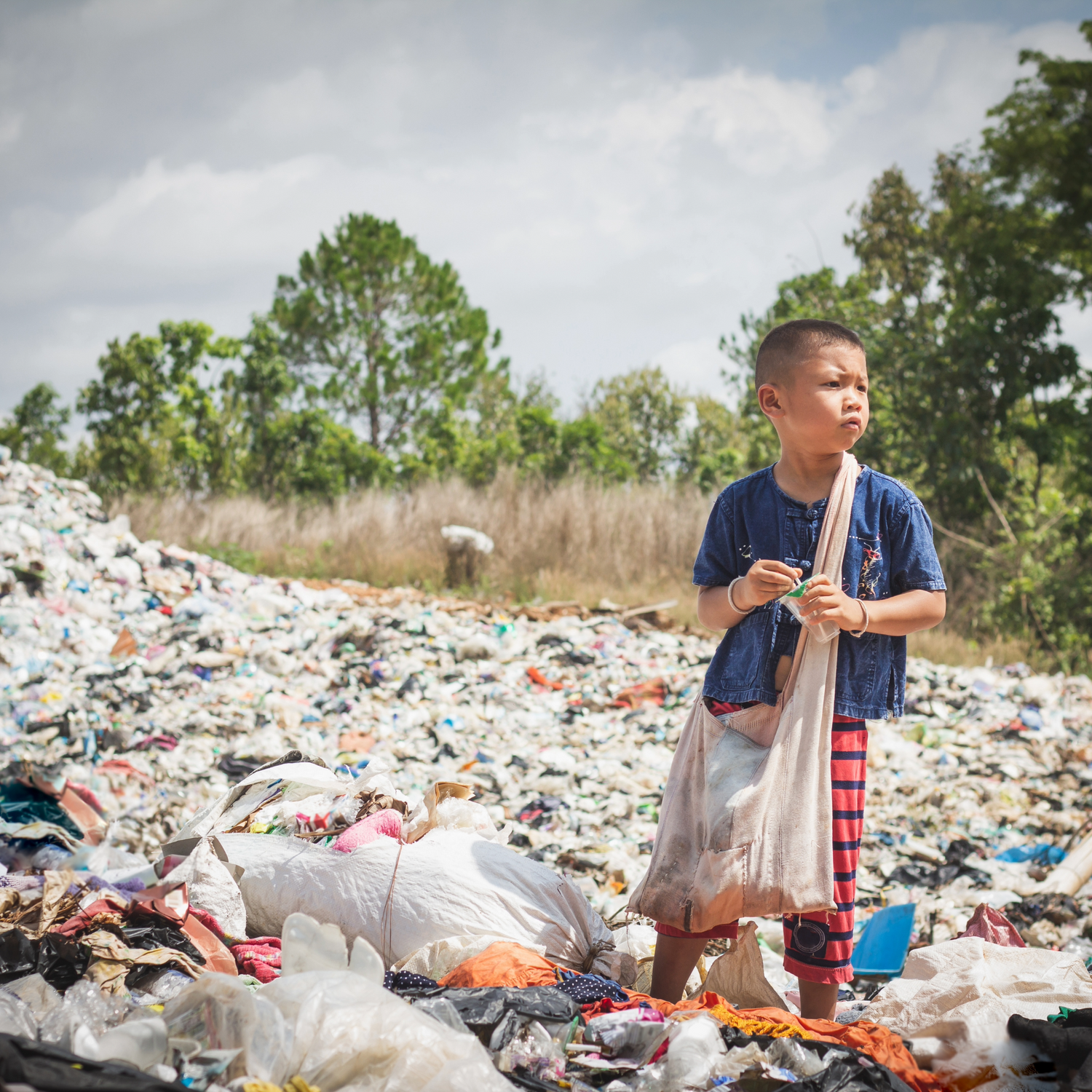A young boy standing on a pile of accumulated plastic film and plastic pollution in his environment looking sad.
